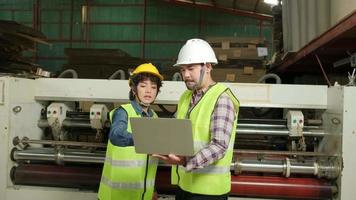 Safety uniform workers and industrial engineers with hardhat use laptop computer to check and control machines. Two professionals work in paper manufacturing factory, maintain production equipment. video