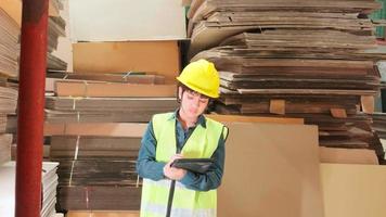 trabajadora con uniforme de seguridad y casco, inspectora supervisora con una tableta, suministro de pedidos de existencias de embalaje en el almacén de la fábrica, montones de fabricación de papel apilado, industria de producción de reciclaje. video