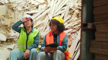 Two female workers and colleagues in safety uniforms and helmets, supervisor teams sit on a pile of used paper for rest, tired from manufacturing work in paper factory, recycling industry occupation. video