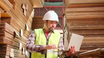 Asian male worker in safety uniform and hard hat, supervisor inspector with laptop for packaging stock order at factory warehouse, piles of stacking paper manufacture, recycling production industry. video