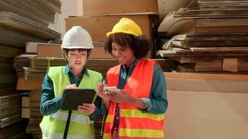 dos trabajadoras y colegas con uniformes de seguridad y verificación de existencias de cascos, controlan la producción en el almacenamiento de la fábrica de almacenes con una gran cantidad de pilas de papel, una amiga trabaja en el negocio de fabricación de reciclaje. video