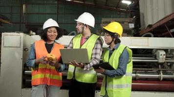 Safety uniform workers and industrial engineers with hardhat use laptop computer to check and control machines. Three professionals work in paper manufacturing factory, maintain production equipment. video