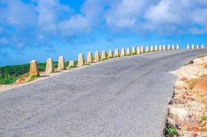 camino asfaltado con cerca de barrera de guardia de piedra que conduce hacia adelante y hacia la derecha, cielo azul nubes blancas foto