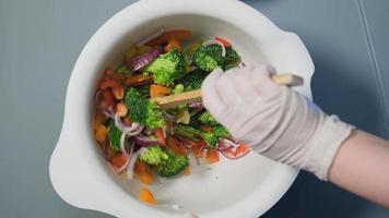 mão na luva mistura salada vegetariana de pimentão brócolis tomate e cebola com colher de pau na tigela closeup extremo video