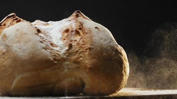 grande pagnotta di pane di grano fresco con farina bianca cade sul tavolo scuro su fondo nero vista estrema vicino al rallentatore