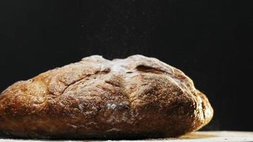Gran hogaza redonda de pan de centeno recién horneado con harina blanca cae sobre la mesa sobre fondo negro cierre extremo cámara lenta