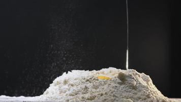 Raw chicken egg with yolk falls down into hole in white flour heap on table on black background extreme closeup slow motion video