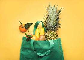 bolsa de compras verde con diferentes frutas, piña, plátanos, naranjas, kiwis y manzanas foto