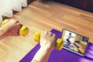 manos de mujeres jóvenes levantando pesas amarillas durante el entrenamiento deportivo en casa, tutoriales de ejercicios de video en la pantalla de la tableta en la alfombra del piso en la sala de estar