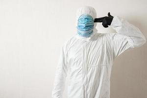 a man in a protective suit hung with medical masks posing against a wall background showing various gestures with his fingers photo