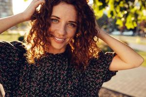 charming curly red-haired girl with freckles in dress poses for the camera in the city center showing different facial emotions photo