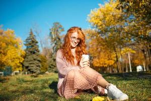 retratos de una encantadora chica pelirroja con gafas y una cara bonita. chica posando en el parque de otoño con un suéter y una falda de color coral. foto