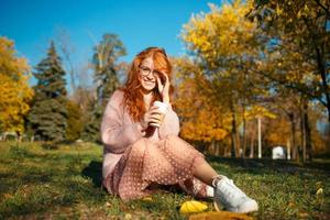 retratos de una encantadora chica pelirroja con gafas y una cara bonita. chica posando en el parque de otoño en un suéter y una falda de color coral. foto