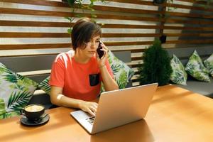 Portrait of confident mature professional woman in glasses, a coral T-shirt sitting on summer terrace in cafe, using laptop computer for work, laughing happily indoors photo