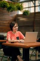 Portrait of confident mature professional woman in glasses, a coral T-shirt sitting on summer terrace in cafe, using laptop computer for work, laughing happily indoors photo