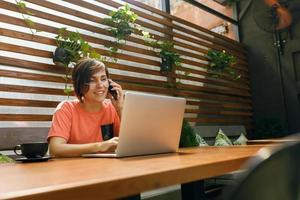 retrato de una mujer profesional madura y segura de sí misma con anteojos, una camiseta de coral sentada en una terraza de verano en un café, usando una computadora portátil para trabajar, riéndose alegremente en el interior foto