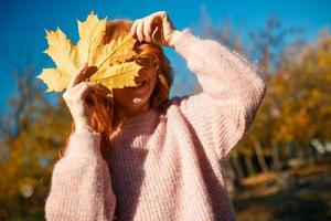 Portraits of a charming red-haired girl with a cute face. Girl posing in autumn park in a sweater and a coral-colored skirt. In the hands of a girl a yellow leaf photo