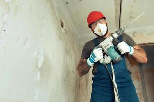a builder with a perforator poses against the background of a wall with empty space from text The builder is dressed in a protective suit and helmet photo