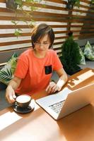 Portrait of confident mature professional woman in glasses, a coral T-shirt sitting on summer terrace in cafe, using laptop computer for work, laughing happily indoors photo