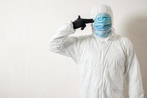 a man in a protective suit hung with medical masks posing against a wall background showing various gestures with his fingers the scientist gestures a pistol to his temple photo