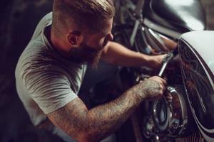 un apuesto hombre barbudo con chaqueta de cuero y gafas de sol está sentado en la motocicleta en el taller de reparación foto