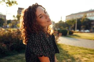 adorable curly red-haired girl with freckles in a dress enjoys good weather sitting on the lawn photo