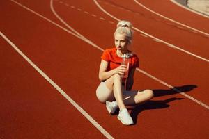 joven y bella rubia se sienta en una pista de jogging con una botella de agua en las manos foto