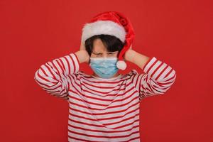 angry kid with medical mask wearing Santa Claus hat with his hands on his head photo
