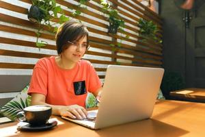 retrato de una mujer profesional madura y segura de sí misma con anteojos, una camiseta de coral sentada en una terraza de verano en un café, usando una computadora portátil para trabajar, riéndose alegremente en el interior foto