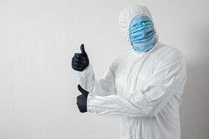 a man in a protective suit hung with medical masks posing against a wall background showing various gestures with his fingers, the scientist shows a thumb up on both hands photo