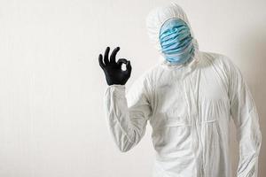 a man in a protective suit hung with medical masks posing against a wall background showing various gestures with his fingers the scientist shows ok photo