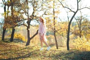 retratos de una encantadora chica pelirroja con una cara linda. chica posando en el parque de otoño con un suéter y una falda de color coral. en manos de una niña una hoja amarilla foto