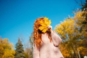 retratos de una encantadora chica pelirroja con una cara linda. chica posando en el parque de otoño con un suéter y una falda de color coral. en manos de una niña una hoja amarilla foto