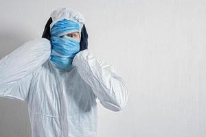 un hombre asustado con un traje protector colgado con máscaras médicas contra una pared blanca. los horrores de la epidemia, el peligro del coronavirus. el científico sostiene su cabeza con horror foto