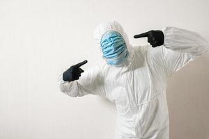 man in a protective suit posing against wall background showing various gestures with his fingers. scientist gestures with his hands to indicate a set of medical masks on his face photo