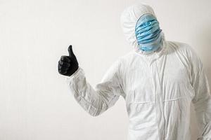 a man in a protective suit hung with medical masks posing against a wall background showing various gestures with his fingers photo