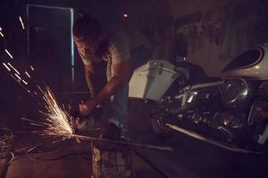 Handsome brutal male with a beard repairing a motorcycle in his garage working with a circular saw. In the garage a lot of sparks and smoke from sawing