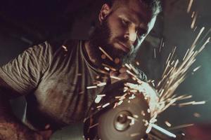 Handsome brutal male with a beard repairing a motorcycle in his garage working with a circular saw. In the garage a lot of sparks and smoke from sawing photo