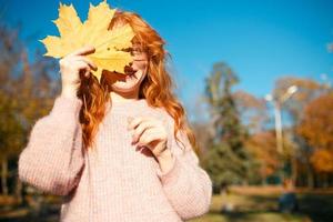 retratos de una encantadora chica pelirroja con una cara linda. chica posando en el parque de otoño con un suéter y una falda de color coral. en manos de una niña una hoja amarilla foto