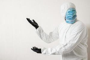 man in a protective suit hung with medical masks posing against wall background showing various gestures with his fingers. the scientist gestures to the wall where the text can be placed photo