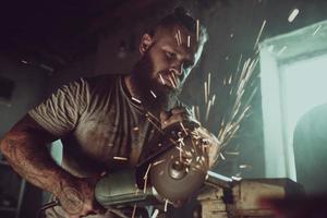 Handsome brutal male with a beard repairing a motorcycle in his garage working with a circular saw. In the garage a lot of sparks and smoke from sawing photo