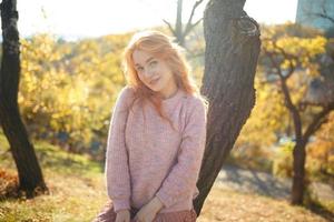 Portraits of a charming red-haired girl with a cute face. Girl posing in autumn park in a sweater and a coral-colored skirt. In the hands of a girl a yellow leaf