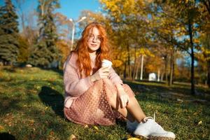 retratos de una encantadora chica pelirroja con gafas y una cara bonita. chica posando en el parque de otoño en un suéter y una falda de color coral. foto