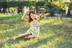 little girl is played by photo camera sitting on grass in park. Doing Selfie and photographing the world around