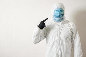 a man in a protective suit hung with medical masks posing against a wall background showing various gestures with his fingers photo