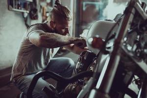 hombre guapo con barba reparando su motocicleta en el garaje. un hombre vestido con jeans y una camiseta foto