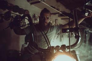 un hombre guapo y brutal con barba sentado en una motocicleta en su garaje, limpiándose las manos y mirando hacia un lado foto