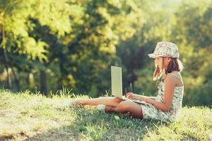 little girl is talking on a laptop while sitting on the grass in the sun. Dressed in a sarafan and hat photo
