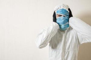 a frightened man in a protective suit hung with medical masks against a white wall. The horrors of the epidemic, the danger of the coronavirus. the scientist holds his head in horror photo