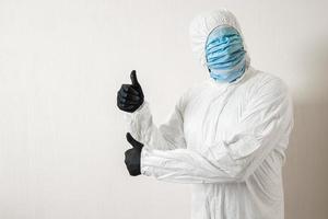 a man in a protective suit hung with medical masks posing against a wall background showing various gestures with his fingers, the scientist shows a thumb up on both hands photo